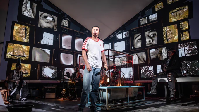 An average height, slim, Afro-Caribbean man wearing a red and white top and jeans stands in front of two large walls of retro tv screens in a V-formation. Each of the tv screens depicts a different image, like a portion of his face. Other figures stand around the area.