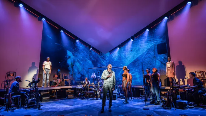 A group of people stand on a wide stage, with light pink walls and ceiling, and walls portraying blurred video images. There are amps and instruments littered around the stage, including a drum kit and guitars. There are also chairs and other props dotted around the stage.