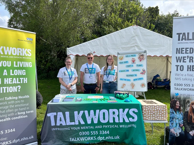 Talkworks stand at Devon County Show. Three members of staff from Talkworks are wearing Talkworks T shirts and standing behind a table with a Talkworks table cloth. On the table are leaflets, pens, stress balls and a buzz wire game. A white board that saysw Win a prize. Test your fight or flight – a normal and natural response to help you cope with stress. Two Talkworks pull up banners are on either side of the table. One says Talkworks are you living with a long-term health condition, the other Talkworks we’re here to help. On the tablecloth 0300 555 3344 talkworks.dpt.nhs.uk 