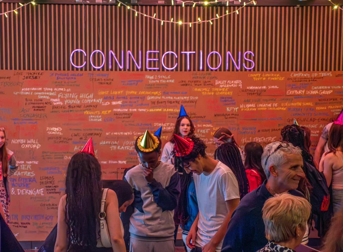 Young people wearing party hats in front of a pink neon sign reading 'Connections'