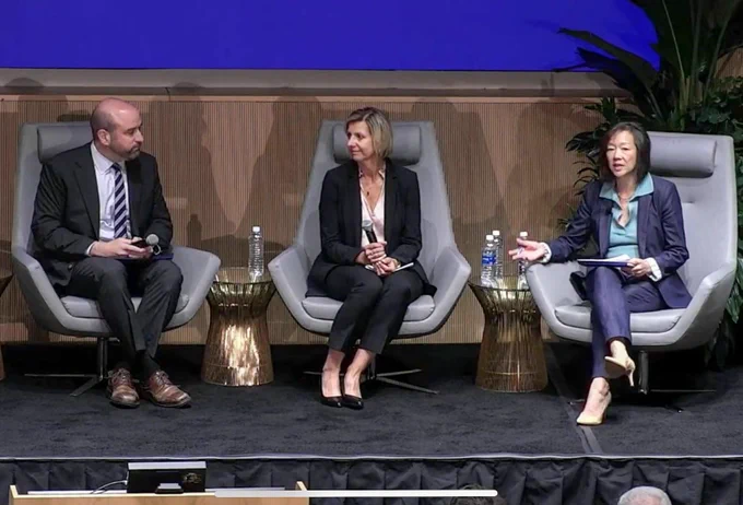 Three individuals in business attire sit on stage in armchairs, engaged in a discussion. The woman on the right gestures while speaking, with two water bottles on a small table to her left.