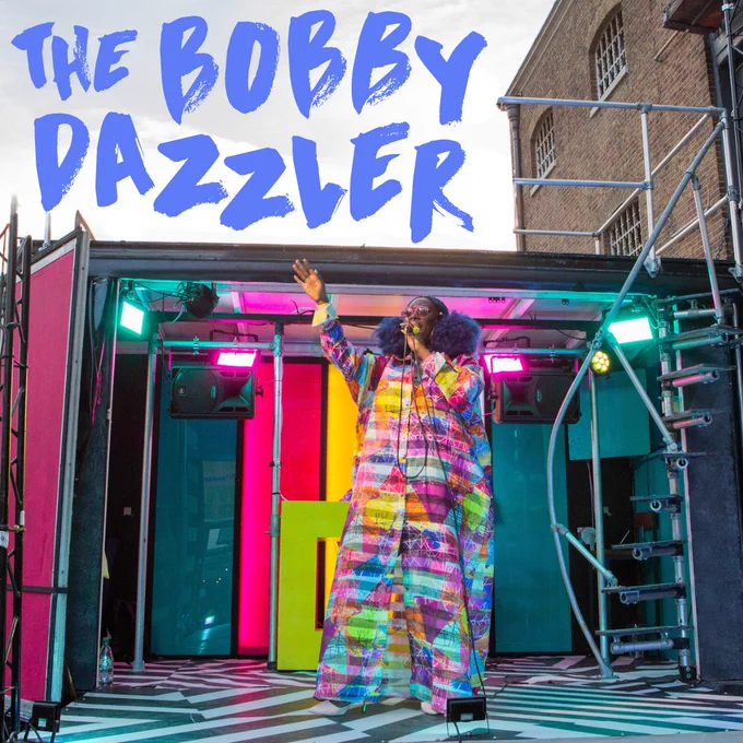 Photo of The Bobby Dazzler. A black and white striped van has the side panel removed to transform the van into a performance stage. Inside the van are multi-coloured panels and lighting. There is a performer on the stage with a microphone and arm raised in the air; they are wearling a bright multi-coloured coat. Above them is the text "The Bobby Dazzler".