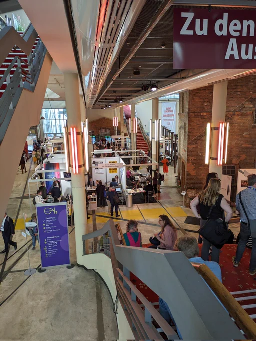 This is a photos taken at the top of a staircase, looking down into the expo hall of Classical Next conference. The expo hall is in a large bright cinema foyer space with large white pillars, a high ceiling and some exposed brick walls. There are expo stalls on the floor, and people are milling around.