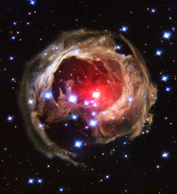 V838 Mon. Large, prominent stars with diffraction spikes are surrounded by a shell of illuminated gas and dust. A central star shines in red, and the others are blue and white. The shining dust around them is light brown.