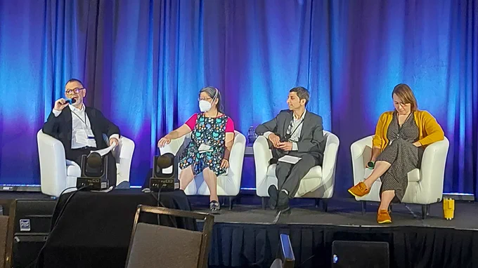 Dr. Alan Katz, a white man with short hair and glasses, speaks into a microphone as the moderator of a panel discussion. Panelists Amy Ma, an asian woman wearing a mask, Dr. Onil Bhattacharyya, a south asian man with short hair, and Dr. Ruth Lavergne, a white woman with long dark hair, sit on chairs on a stage. 
