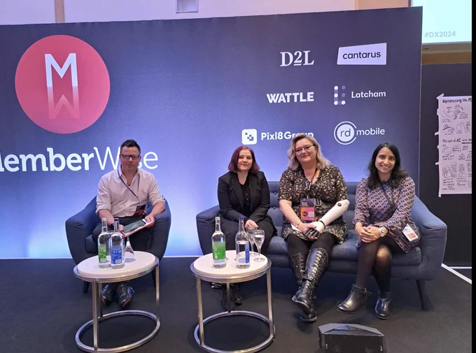 Three women on a blue sofa and a man on a blue chair smile at the camera in front of a conference backdrop. There are lots of logos on the backdrop and the central text reads Memberwise