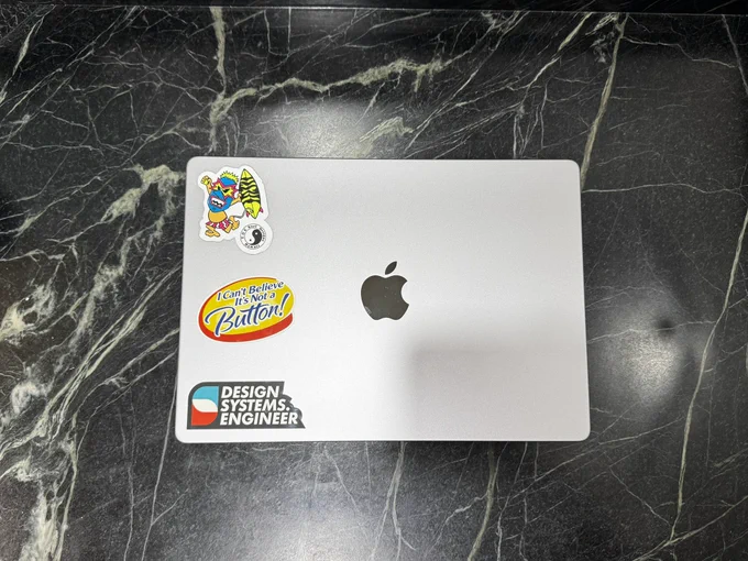 A MacBook with 3 stickers: a T & C Surf Tiki Man, I can’t believe it’s not a button, and a design systems engineer logo.