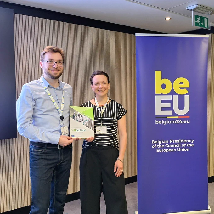 A man and a woman standing in a conference room next to a banner which shows the following text: "Belgian Presidency of the Council of the European Union". They hold the PROACTIVE final brochure in their hands.