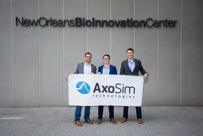Three individuals stand in front of the New Orleans BioInnovation Center holding a banner for AxoSim Technologies.