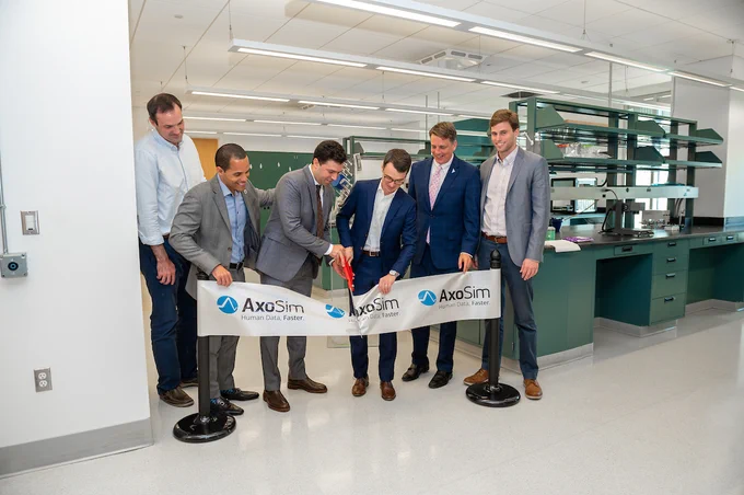 Five individuals participate in a ribbon-cutting ceremony inside a laboratory, representing the company AxoSim. They stand behind a banner featuring the company’s logo. The setting includes laboratory benches and equipment in the background.