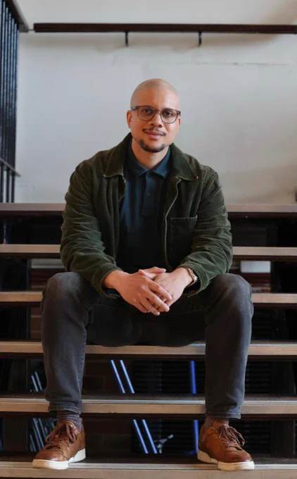 Writer Nathaniel Price is sat on a flight of stairs at Chapter Arts Centre. He has his hands clasped in front of him and smiles towards the camera