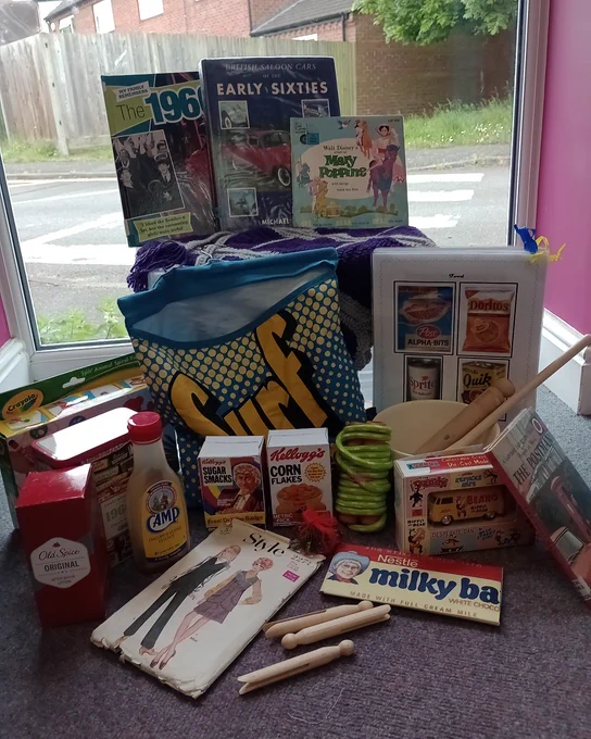 60's reminiscence box, magazines, books, milky bar, clothes pegs, clothes pattern, cereal boxes, rolling pin, skipping rope, cooking book, biscuit box, surf bag, games, bus in front of a window