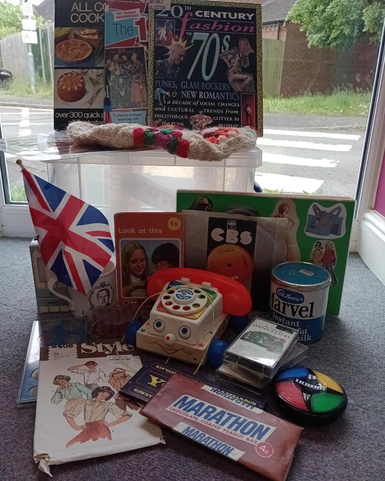 70's reminiscence box, British flag, toy telephone, marvel powder milk tin, marathon chocolate, clothes pattern, games, Yorkie bar chocolate, crochet bag, books, CBS record/vinyl, look at this book, Style magazine in front of a window.