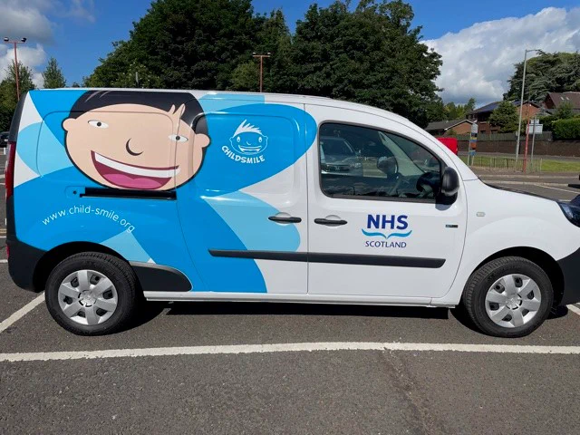 The blue Childsmile van sitting in  a car park with some trees in the background and road signs. 