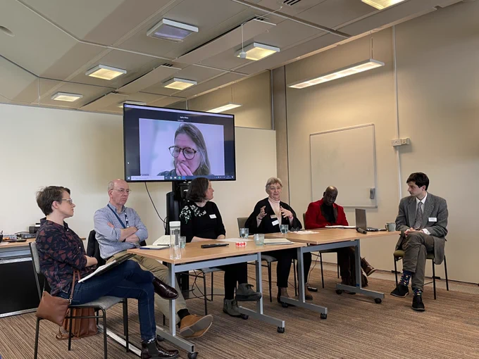 Six people sit on one side of a conference table with one more person appearing live on a television screen behind them.