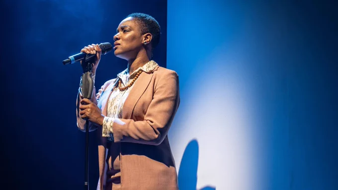 A Black woman of average height with short hair wearing a peach coloured suit sings into a microphone