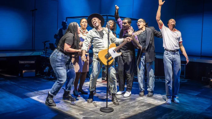 A group of people sing together. In the centre, one figure, wearing a boiler suit and a wide-brimmed hat, and playing an electric guitar, sings into a microphone. The others stand around him, singing along.