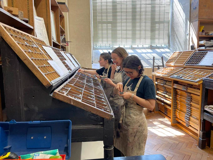 Three trainees stand infront of cabinets full of type, they are typesetting their names by picking up letters and placing them in their supports in the correct manner. 