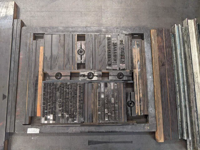 A photograph of a typeset printing block, with supports and re-enforcements to ensure the type remained tightly set in place. The type is made from lead, as is the frame and some supports, some are made from wood.