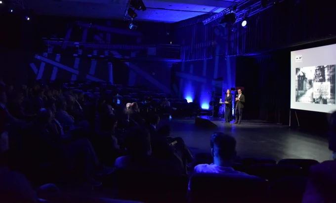 Audience attending a presentation in a dimly lit auditorium with a speaker standing next to a large screen displaying a slide.

