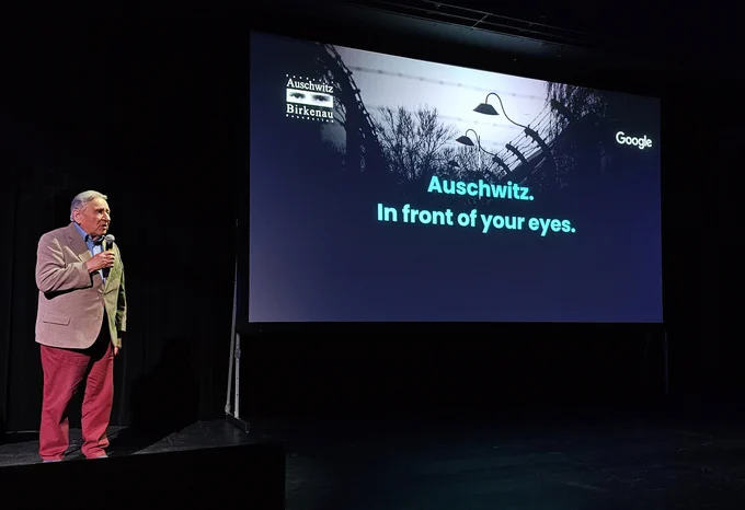 An elderly person - Auschwitz Survivor Bogdan Bartnikowski - stands onstage holding a microphone, beside a large screen displaying the Google logo and an image of a barbed wire fence under a dark sky. The screen reads "Auschwitz. In front of your eyes."