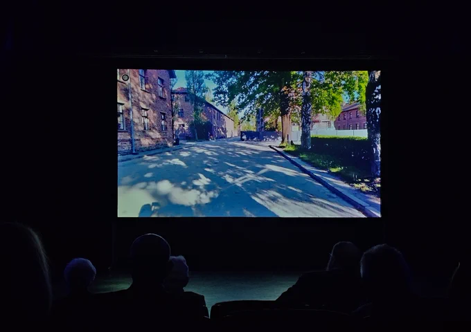 Audience watching an online guided tour of the Auschwitz Memorial. The street of Auschwitz I site is visible in the screen. 
