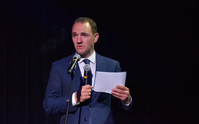 Rowan Barnett delivers a speech at a podium with a microphone, holding a sheet of paper, set against a dark stage background.
