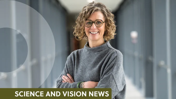 A woman with short curly hair. She is wearing a glasses and is smiling at the camera. She is standing with her arms folded and is wearing a grey jumper. 