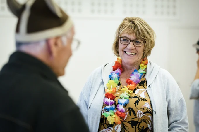 An adult with bobbed blonde hair wearing a brightly coloured flower garland is smiling at another adult with their back to the camera, who is wearing a Viking helmet.