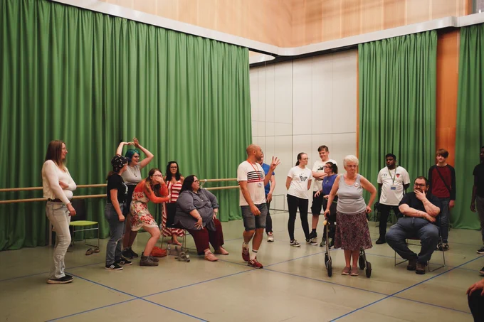 A rehearsal room with green curtains and a grey floor separated into squares with blue tape. James is in the middle of a group of people, some are in various animated shapes either sitting or standing. James is wearing a white tshirt and shorts and holding up four fingers to the group. 