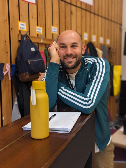 James Blakey, a white man with a brown short beard is smiling at the camera, resting his head on one hand. He's wearing a teal adidas tracksuit top and there is a yellow water bottle in front of him.