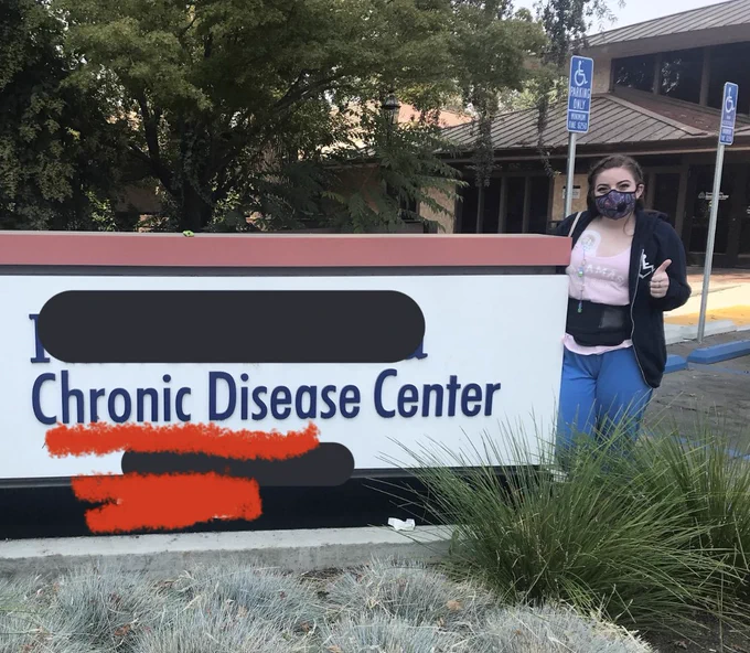 A large “Chronic Disease Center” sign in front of a building. A young adult Moe stands to the right of the sign, giving a thumbs up. Her portacath is visible on her chest and she is wearing a back brace.