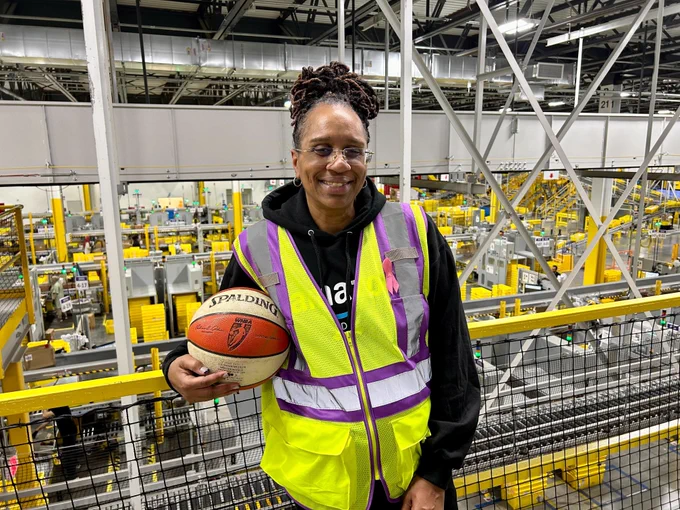 Dena Head, Amazon HR Manager, standing in an Amazon fulfillment center, wearing a yellow safety vest and holding a basketball.
