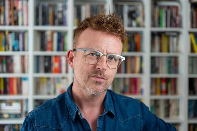 Steve Cross in a blue shirt looking thoughtful in front of an out-of-focus bookcase.