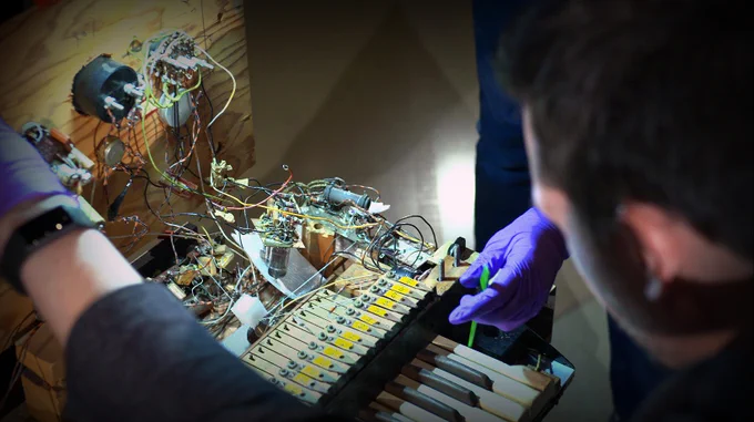 Over-the-shoulder shot of curator and conservator peering under the hood of the Electronic Sackbut synthesizer. Internal wiring and circuitry behind keyboard is exposed.