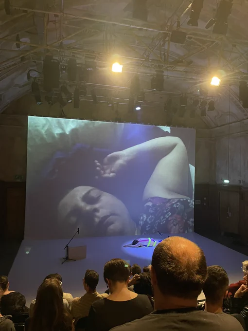 A projection screen shows a woman lying down with her eyes closed and arm resting above her head. On the stage there is a white dance floor with a woman lying face down wearing coloured body tape. An audience watches on.