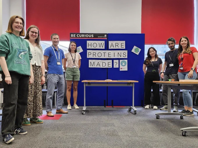 A group of people stand beside board labelled 'How are proteins made?'.