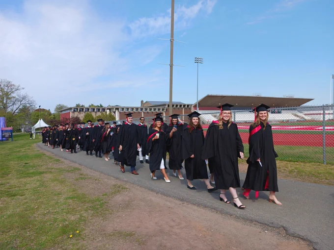 A large group of graduates March toward their ceremony under a bright blue sky. They are wearing black robes, mortar caps, and the red satin trimmed hoods of the Acadia BBA program. Raymond field is in the background.
