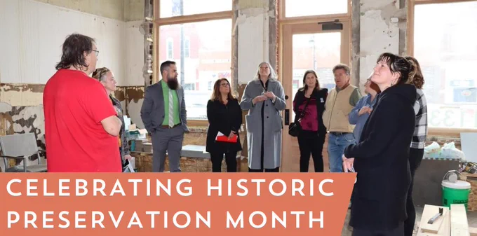 A group of people attentively listen to a speaker during a tour inside a large, unfinished room with exposed brick walls and large windows. Text reads "celebrating historic preservation month"