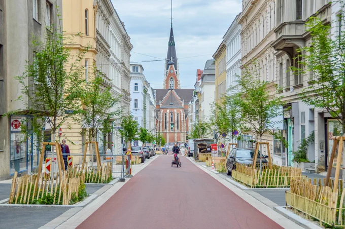 People in casual spring attire cycle on Argentinierstraße in Vienna—a red asphalt “bicycle street” lined with four-storey buildings, trees and plants, and wooden benches. 