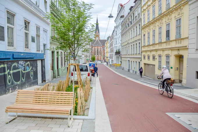 People in casual spring attire cycle on Argentinierstraße in Vienna—a red asphalt “bicycle street” lined with four-storey buildings, trees and plants, and wooden benches. 