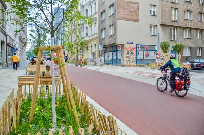 People in casual spring attire cycle on Argentinierstraße in Vienna—a red asphalt “bicycle street” lined with four-storey buildings, trees and plants, and wooden benches. 