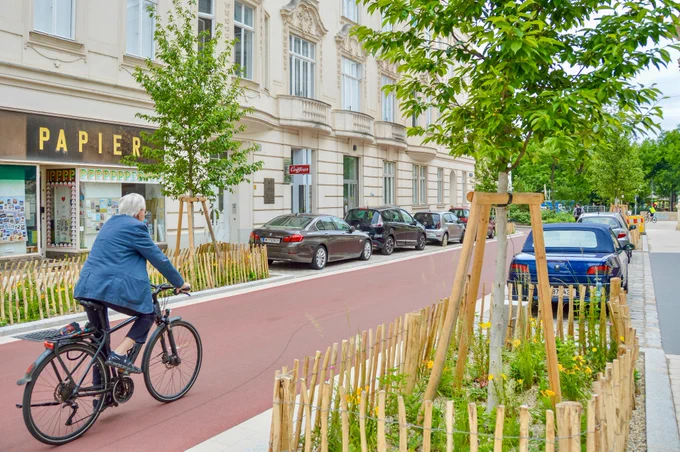 People in casual spring attire cycle on Argentinierstraße in Vienna—a red asphalt “bicycle street” lined with four-storey buildings, trees and plants, and wooden benches. 