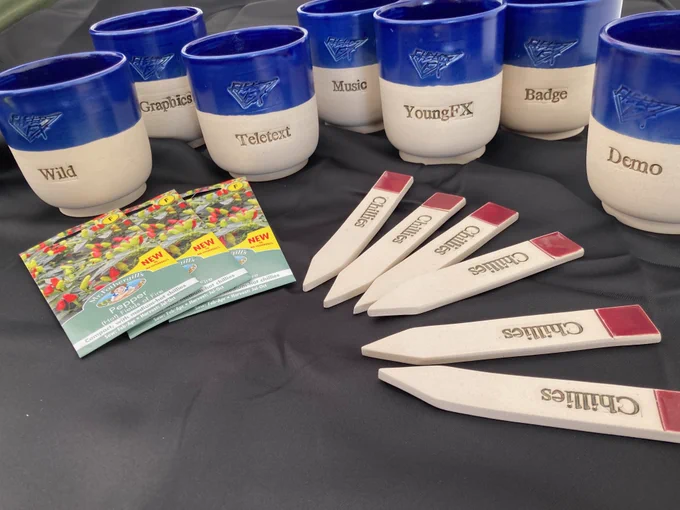 Seven pots, with blue tops and white bases, with Field-FX logos embossed and the words "wild" "graphics" "teletext" "music" "YoungFX" "Badge" "Demo" on them. In the foreground there are chilli seeds and matching "Chillies" plant labels made in the same pottery as the pots, with red tops.