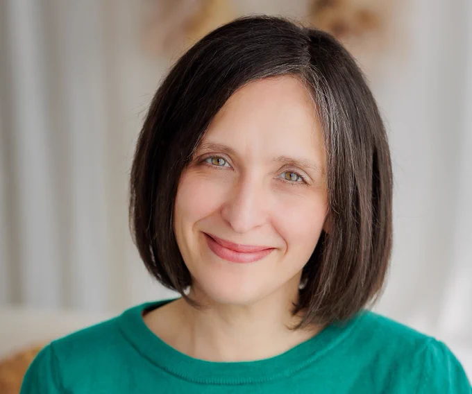 Susanna is a white woman with straight dark shoulder length hair. She's wearing a green jumper and smiling to the camera in a head and shoulders shot.