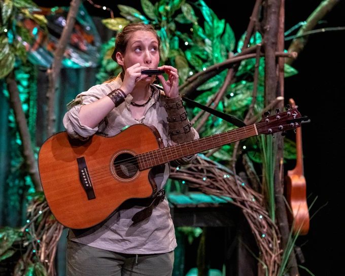 A person on a set that looks like a den in a forest. They are holding an acoustic guitar and playing a mouth organ.