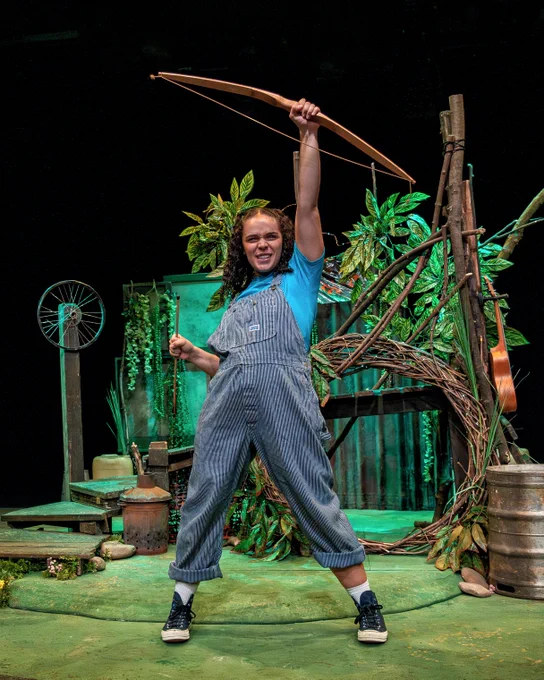 A person wearing denim dungarees and a blue t-shirt holding a wooden bow and arrow. They are on a set that looks like a den in a forest.