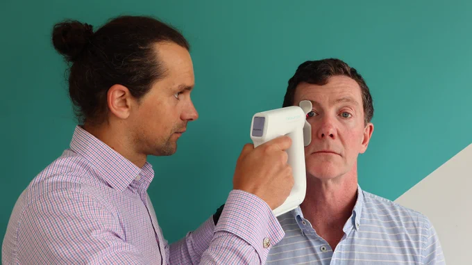 Two people are pictured against a turquoise and white wall. One person holds up a small handheld eye scanning machine to the other person's right eye. The second person stares again in a relaxed way. 
