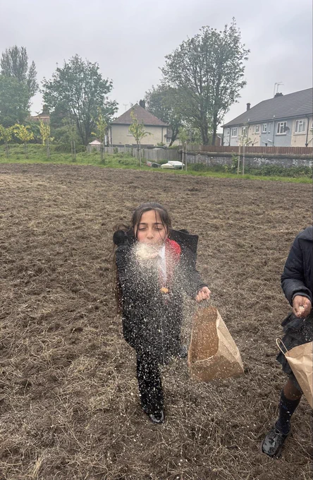 A young person standing in a ploughed field blowing a handful of seeds onto the ground.