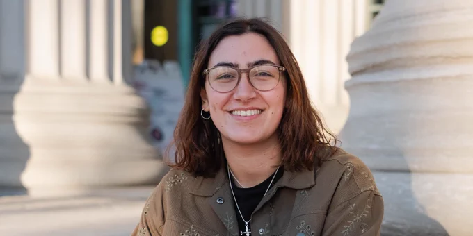 Person smiling, seated on a step in front of a building with columns, wearing glasses, a brown jacket, and jeans.